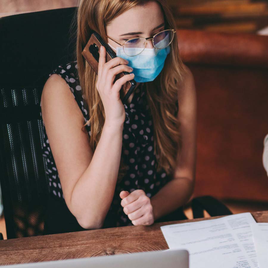 A woman sitting at desk wearing a mask while on the phone