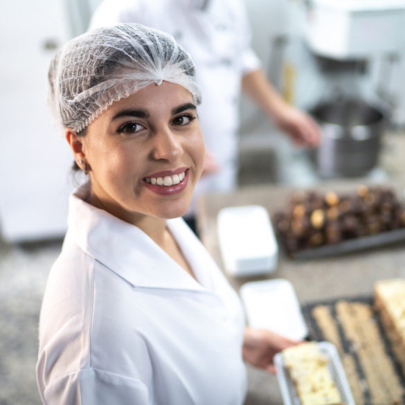 woman wearing hair net smiling at camera with food on tabel behind her